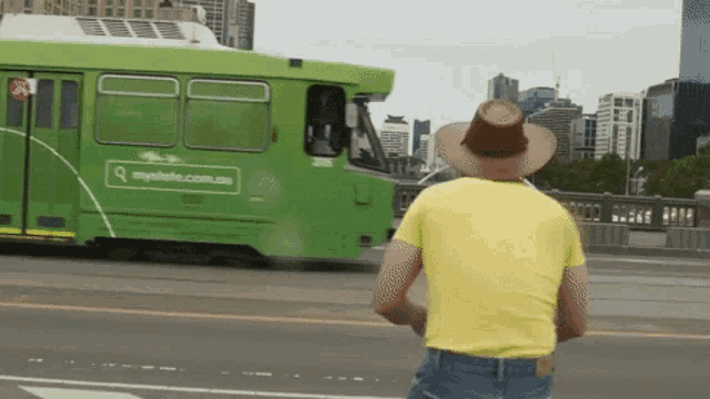 a man wearing a cowboy hat stands in front of a green bus that says mystate.com.au on the side