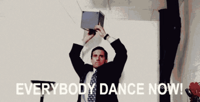 a man in a suit and tie is holding a box over his head with the words `` everybody dance now '' written below him .