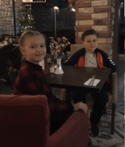 a boy and a girl sit at a table with a vase of flowers on it