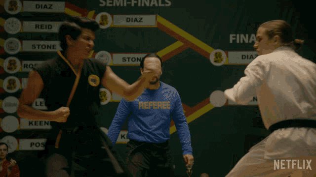 a man in a blue shirt that says referee on it watches two karate fighters