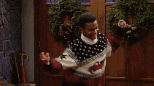a man wearing a christmas sweater is standing in front of a door with a wreath on it .