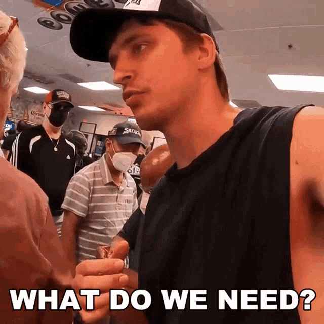 a man wearing a black tank top and a black hat stands in front of a group of people and says " what do we need "