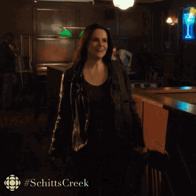 a woman in a leather jacket stands in front of a bar with #schitts creek on the bottom right