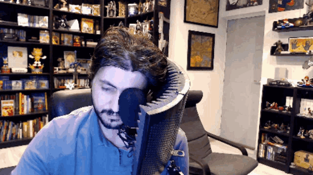 a man in a blue shirt is sitting in front of a microphone in a room full of bookshelves
