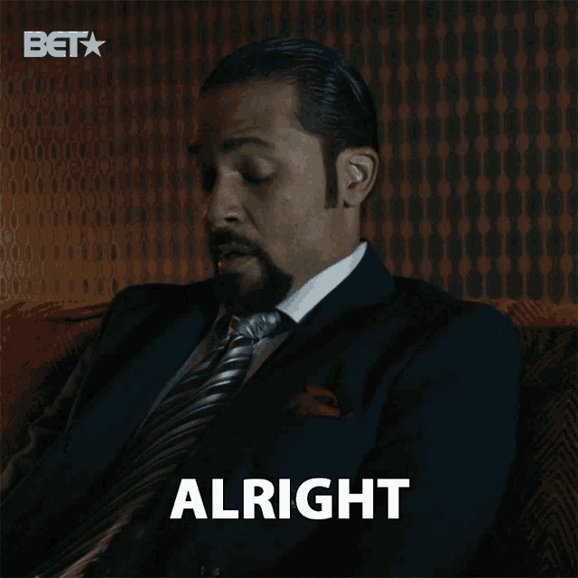 a man in a suit and tie is sitting on a couch and says alright