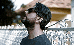 a man with a beard wearing sunglasses is standing in front of a fence .