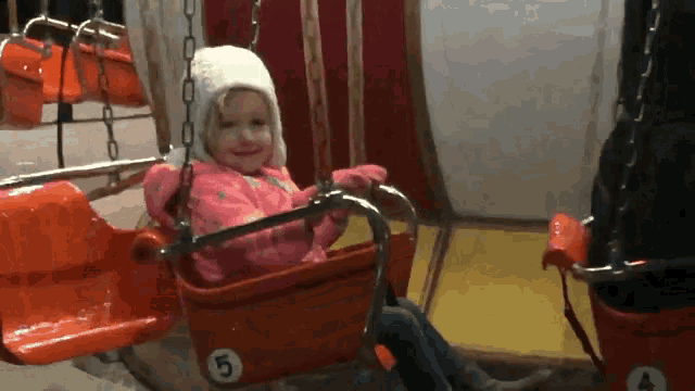 a little girl in a pink jacket is sitting in a red swing with the number 5 on it