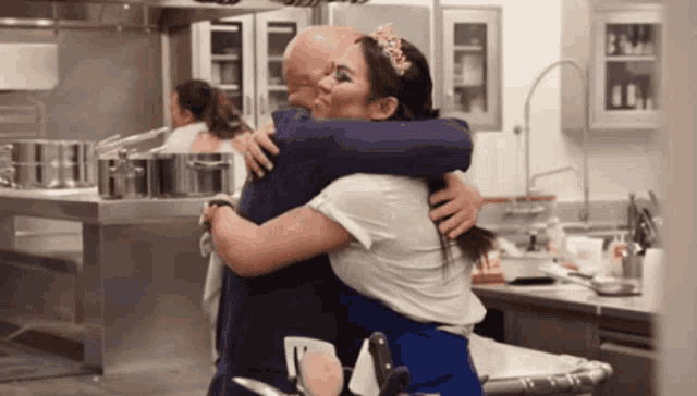 a man and woman hugging in a kitchen with pots and pans behind them