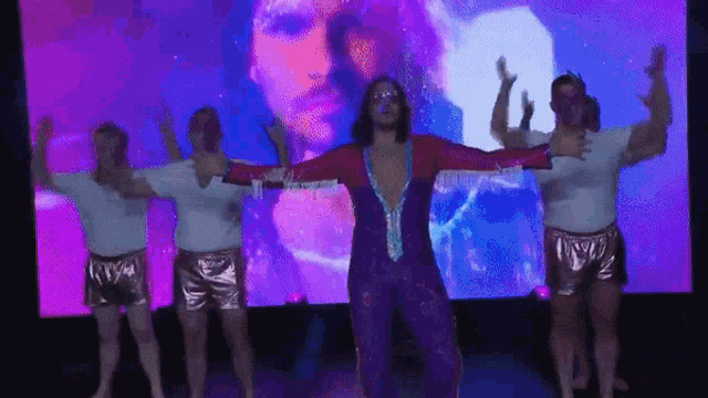 a group of men are dancing in front of a large screen with a man 's face projected on it