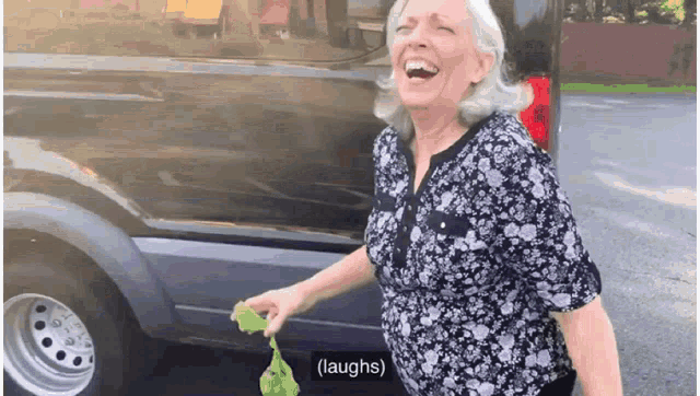 an elderly woman is laughing in front of a van while holding a green bag .