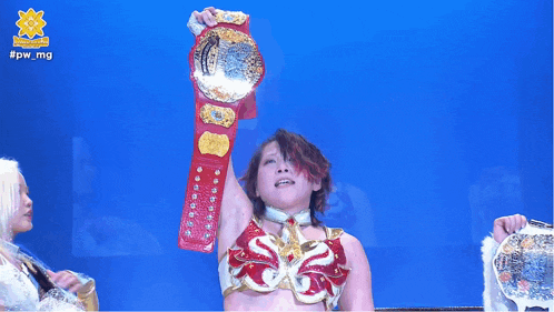 a female wrestler holds up her championship belt in front of a blue background that says #pwmg