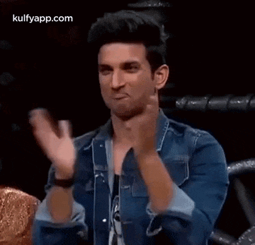 a man in a denim jacket is clapping his hands while sitting in front of a microphone .