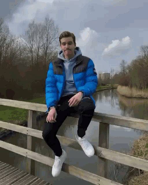 a man in a blue jacket is sitting on a wooden bridge over a river