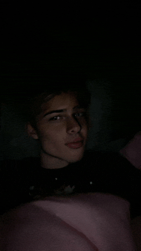 a young man is laying in bed with a pink blanket and a black shirt on .
