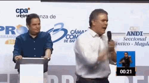 a man stands at a podium with a microphone while another man stands behind him