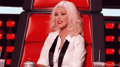 christina aguilera is sitting in a red chair on the voice with a drink in her hand .