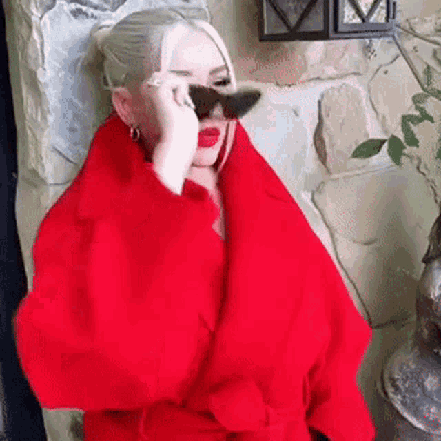 a woman wearing a red coat and sunglasses is covering her face with her hand .