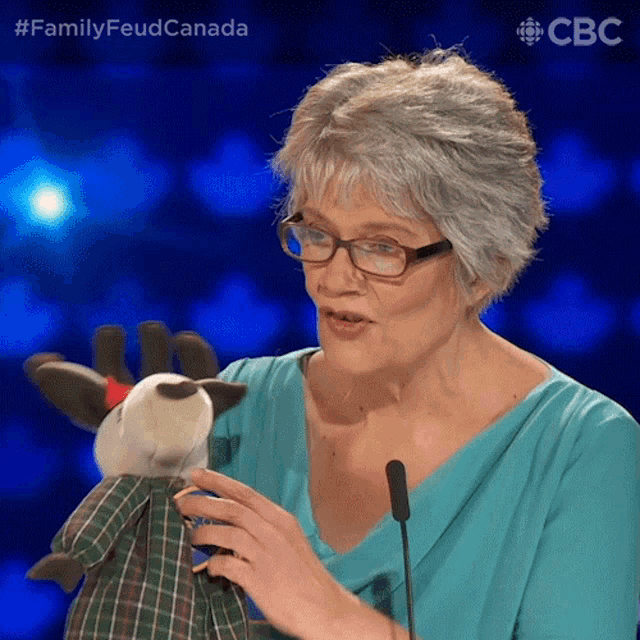 a woman holds a stuffed reindeer in front of a microphone with the hashtag #familyfeudcanada on the bottom