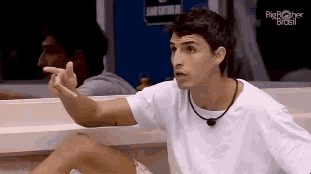 a man in a white t-shirt is sitting in front of a mirror and pointing at something .