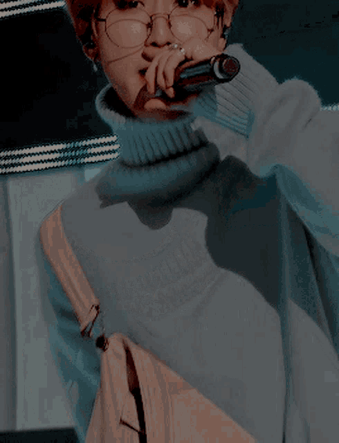 a person wearing glasses and a turtleneck sweater holds a microphone to their mouth