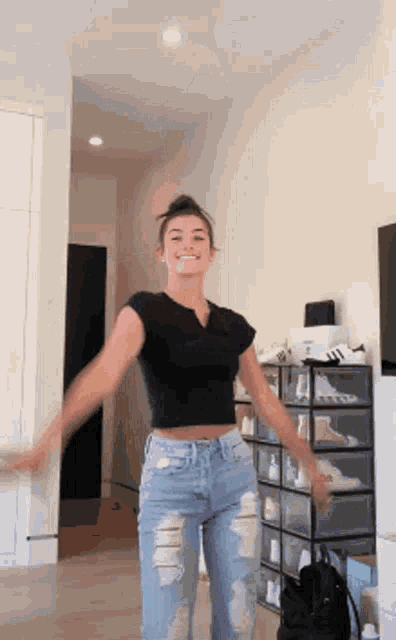 a woman in a black crop top and ripped jeans is dancing in a room with a shoe rack in the background .