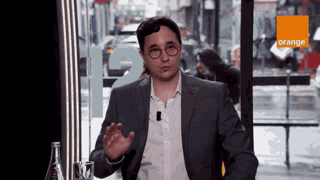 a man in a suit and glasses is talking in front of a sign that says orange