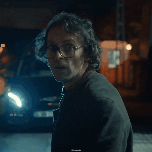 a man with curly hair wearing glasses and a black jacket is looking at the camera with a blue light behind him