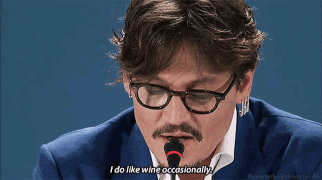 a man with glasses and a beard is speaking into a microphone and says " i do like wine occasionally "