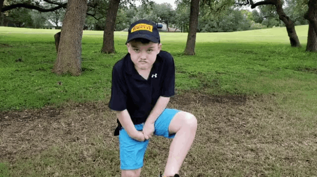 a young boy wearing a black shirt and blue shorts is squatting down in a park