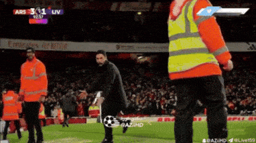 a man in an orange vest stands on a soccer field in front of an emirates fly better advertisement