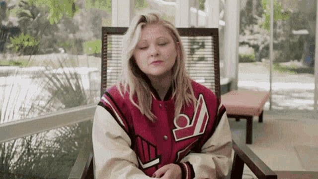 a woman in a red and white varsity jacket is sitting in a chair with her eyes closed .