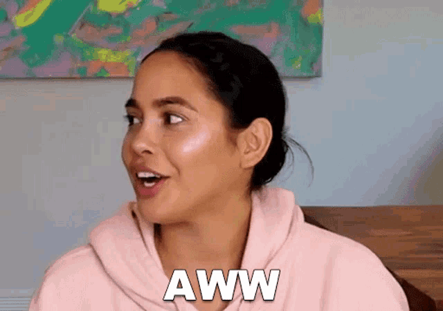 a woman in a pink hoodie is sitting at a table with her mouth open and the word aww written on her face .