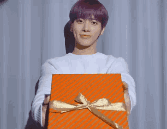 a young man with purple hair is holding a gift box with a gold bow .