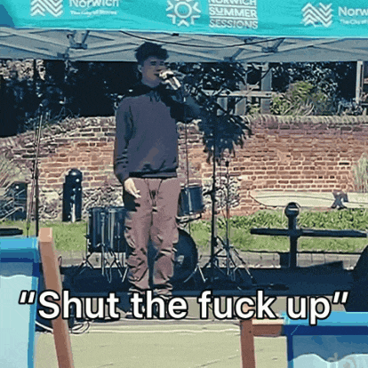 a man singing into a microphone with the words " shut the fuck up " written above him