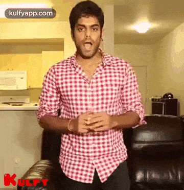a man in a plaid shirt is standing in front of a couch and making a funny face .