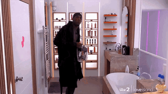 a man in a bathrobe is standing in a bathroom with itv 2 i love island written on the wall