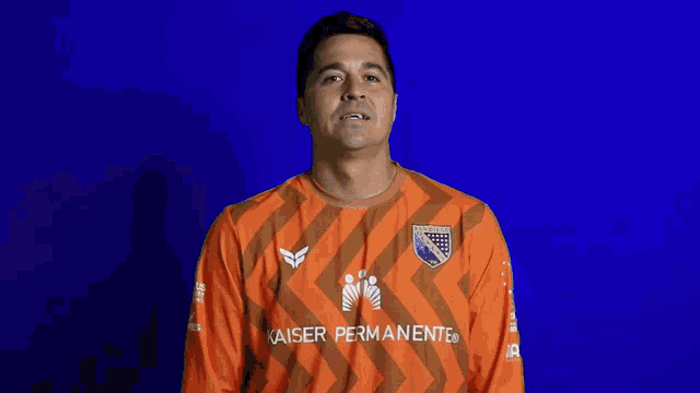 a man wearing an orange kaiser permanente shirt stands in front of a blue background