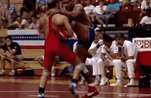 a wrestler in a red singlet is jumping over another wrestler in a blue singlet during a match
