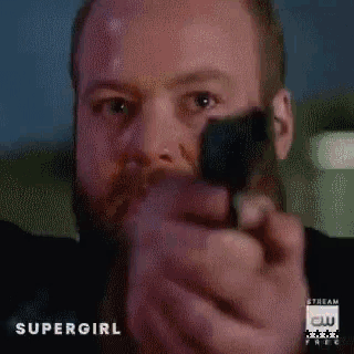 a man with a beard is pointing a gun at the camera with a supergirl logo in the background