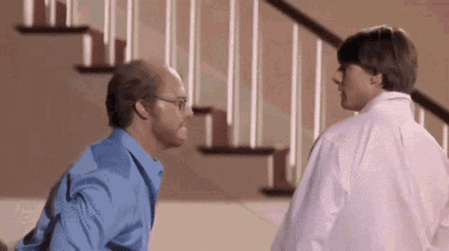 two men are standing next to each other in front of stairs and talking to each other .