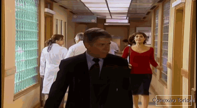 a man in a suit and tie walks down a hospital hallway with a woman in a red shirt