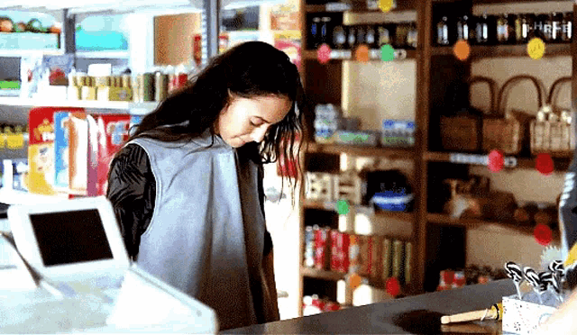 a woman in a grey vest is standing at a counter in a store looking at something
