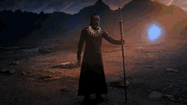 a man in a black robe holds a spear in a desert