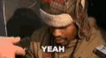 a man wearing a hat and a scarf is talking into a microphone and saying yeah .