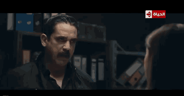 a man with a mustache is talking to a woman in a dark room with a sign that says الحياة