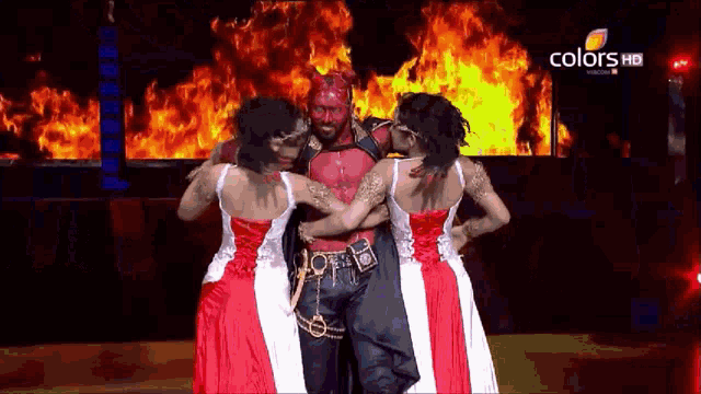 a man with red paint on his face is surrounded by two women on a stage with flames in the background and the words colors hd visible
