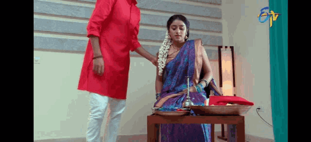 a man in a red shirt is standing next to a woman in a purple saree sitting on a table .