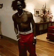 a shirtless man is standing in a living room wearing red shorts and a necklace .