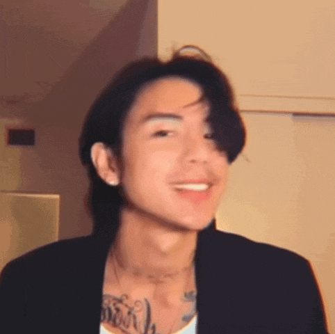 a young man with long hair and a tattoo on his chest is smiling and wearing a black jacket .
