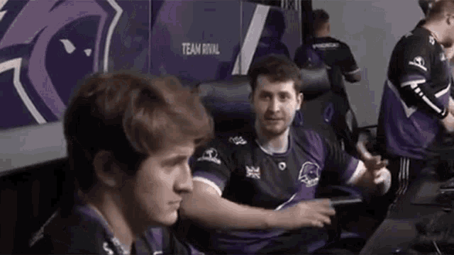 a man in a purple shirt is sitting in front of a team rival sign
